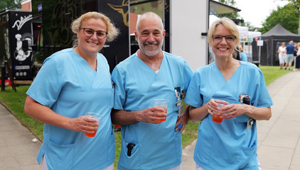 Drei Mitarbeitende in Funktionskleidung halten Getränke in den Händen und lächeln in die Kamera beim Sommerfest 2026 auf dem Gesundheitscampus Volksdorf des Evangelischen Amalie Sieveking Krankenhauses, der Residenz am Wiesenkamp, des Feierabendhauses Volksdorf und der Albertinen Kindertagesstätte Volksdorf. 