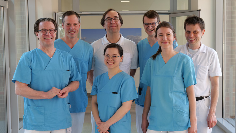 Das Team der Interventionellen AV-Klappen- und Occludertherapie im Albertinen Herz- und Gefäßzentrum steht in weißer und blauer Funktionskleidung auf dem Stationsgang und blickt in die Kamera im Albertinen Krankenhaus in Hamburg