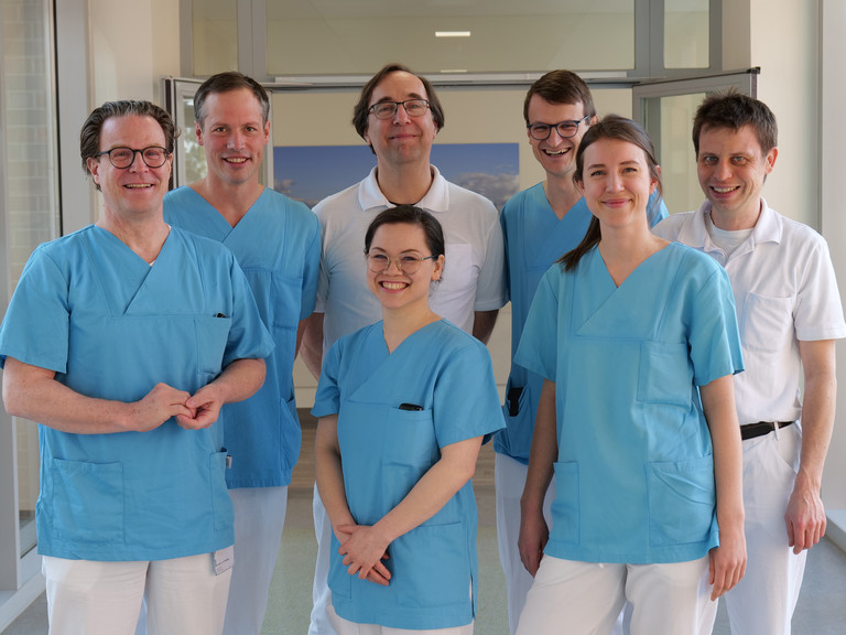 Das Team Des Mitralklappen-Zentrums des Albertinen Herz- und Gefäßzentrums am Standort Hamburg Schnelsen lächelt auf einem Flur in Arztkleidung in die Kamera