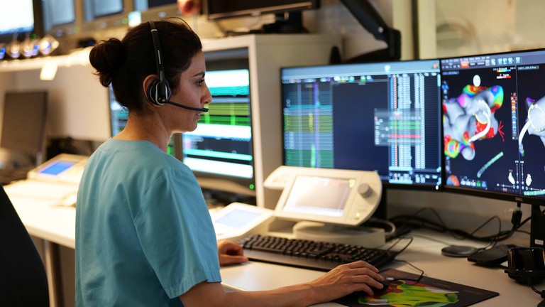 Eine weibliche Fachperson mit Headset und grüner Funktionskleidung sitzt in einem Überwachungsraum während einer elektrophysiologischen Untersuchung im Albertinen Herz- und Gefäßzentrum und blickt auf mehrere Monitore