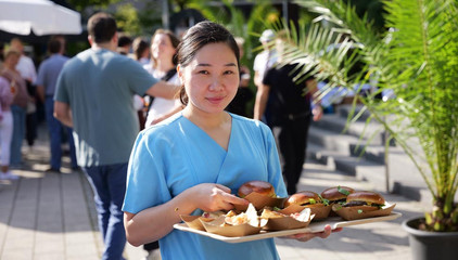 Eine Pflegekraft in blauer Funktionskleidung trägt ein Tablett mit Burger und Pommes beim Albertinen-Sommerfest für die Mitarbeitenden aus den unterschiedlichen Einrichtungen der Immanuel Albertinen Diakonie am Standort Schnelsen auf dem Platz vor der Kirche am Albertinen Krankenhaus.