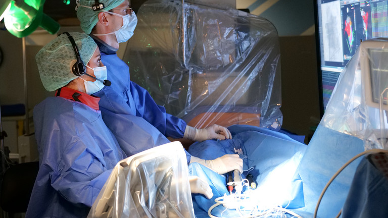 Eine Operateurin und ein Operateur in blauer Schutzkleidung blicken bei einer elektrophysiologischen Prozedur in einem Operationssaal mit medizischen Geräten auf einen Monitor über dem Operationsfeld im Albertinen Herz- und Gefäßzentrum