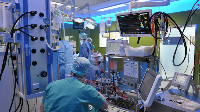 Ein Kardiotechniker sitzt vor einer Reihe medizinischer Gerätschaften mit einem Überwachungsmonitor während einer Operation in einem blau erleuchteten Operationssaal im Albertinen Herz- und Gefäßzentrum in Hamburg