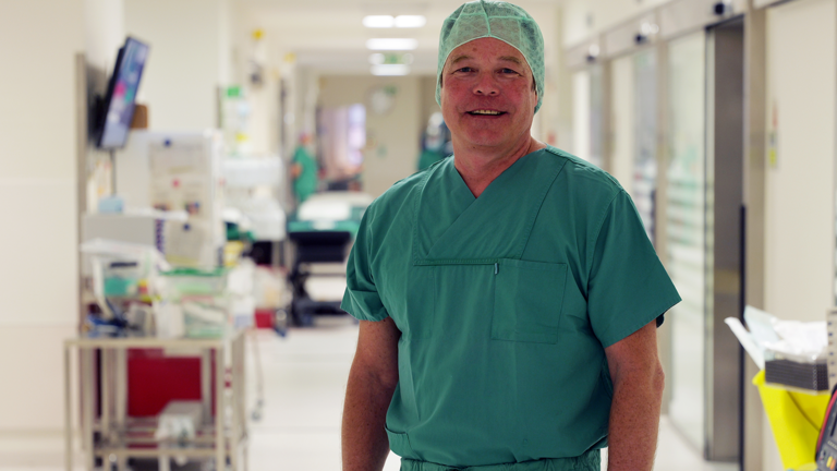 Dr. Ralf Bader, Leitender Oberarzt in der Klinik für Herzchirurgie im Albertinen Krankenhaus, blickt in grüner OP-Kleidung mit Kopfhaube in einem Krankenhausflur in die Kamera und war zu Gast im Podcast von Abendblatt und freigemeinnützigen Krankenhäusern "Hamburger Klinikhelden" zum Thema Bypass-Operationen.