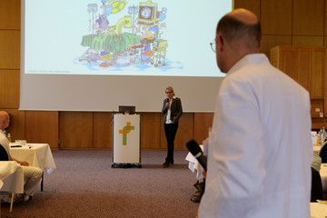 Prof. Dr. med. Ernst von Dobschütz, Chefarzt Zentrum für Endokrine Chirurgie und Ärztlicher Direktor, steht im Vortragsraum und blickt in Richtung der Rednerin auf dem Podium und einer über den Beamer gezeigten Karikatur bei der Fachveranstaltung 11. Amalie Tag der Hausärztinnen und Hausärzte im Evangelischen Amalie Sieveking Krankenhaus in Hamburg-Volksdorf.